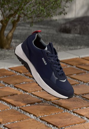 Granatowy sneaker HUGO z białą podeszwą i czarnym bieżnikiem, prezentowany nad drewnianymi kwadratowymi płytkami z małymi białymi kamykami między nimi na zewnątrz.