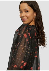 Schwarze durchsichtige Bluse mit plissiertem Design, versehen mit rot-gelben Blumenmustern, hohem Kragen und Rüschelementen an den Schultern.