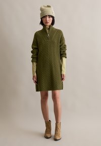 Robe en tricot vert olive avec un motif en chevrons, col zippé quart et manches oversize, portée sur une couche de base jaune clair.