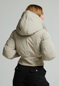 Pufferjack in lichtbeige met een grote capuchon, getailleerde taille en ritsluiting. Heeft gestikte details en merkaanduidingen.