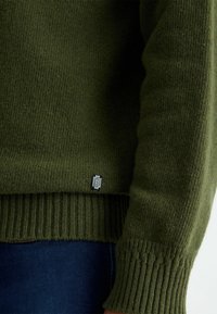 Pull vert en maille avec poignets et ourlet côtelés. Comprend une petite étiquette texturée avec un logo en bas à gauche. Tissu doux de poids moyen.