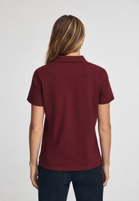 Burgundy pikétröja med krage, korta ärmar, strukturerad tyg. Baksidan har en rak nederkant och minimal detaljering.