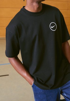 Personne portant un T-shirt noir Nike avec un logo circulaire blanc sur la poitrine et un jean bleu, debout à l'intérieur, une main dans la poche.
