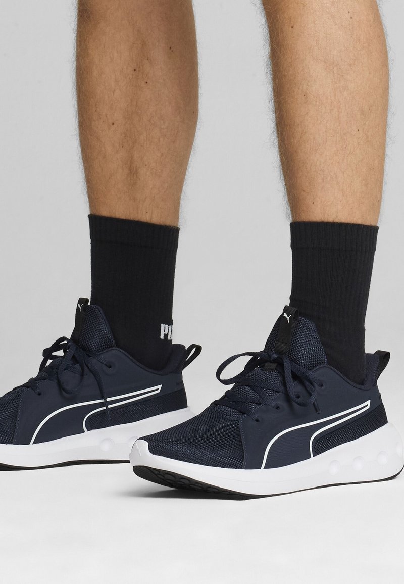 Jambes portant des chaussettes noires Puma et des baskets Puma bleu marine avec semelles blanches sur un fond gris uni.