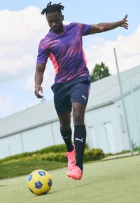 Camiseta desportiva da Puma em roxo com detalhes em rosa, combinada com calções azul-marinho e meias pretas. O jogador está a chutar uma bola de futebol amarela num campo verde.