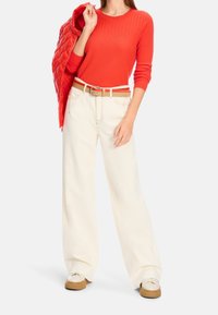 Haut rouge à manches longues côtelé, pantalon large crème, et ceinture beige. Le mannequin tient une veste rouge matelassée. Baskets blanches avec semelles beiges.