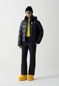 Black puffer jacket with a hood, worn over a yellow top, paired with dark pants and yellow snow boots. Accessories include a black beanie and sunglasses.