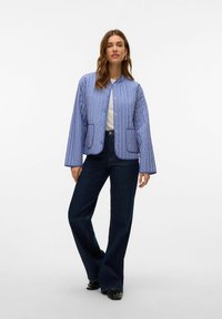 Giacca leggera a righe blu e bianche con chiusura a bottone, due tasche frontali e maniche larghe, abbinata a jeans svasati scuri.