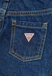 Tessuto denim di colore blu scuro con una tasca posteriore caratterizzata da una patch triangolare bianca con il logo "GUESS" rosso e nero e cuciture gialle.