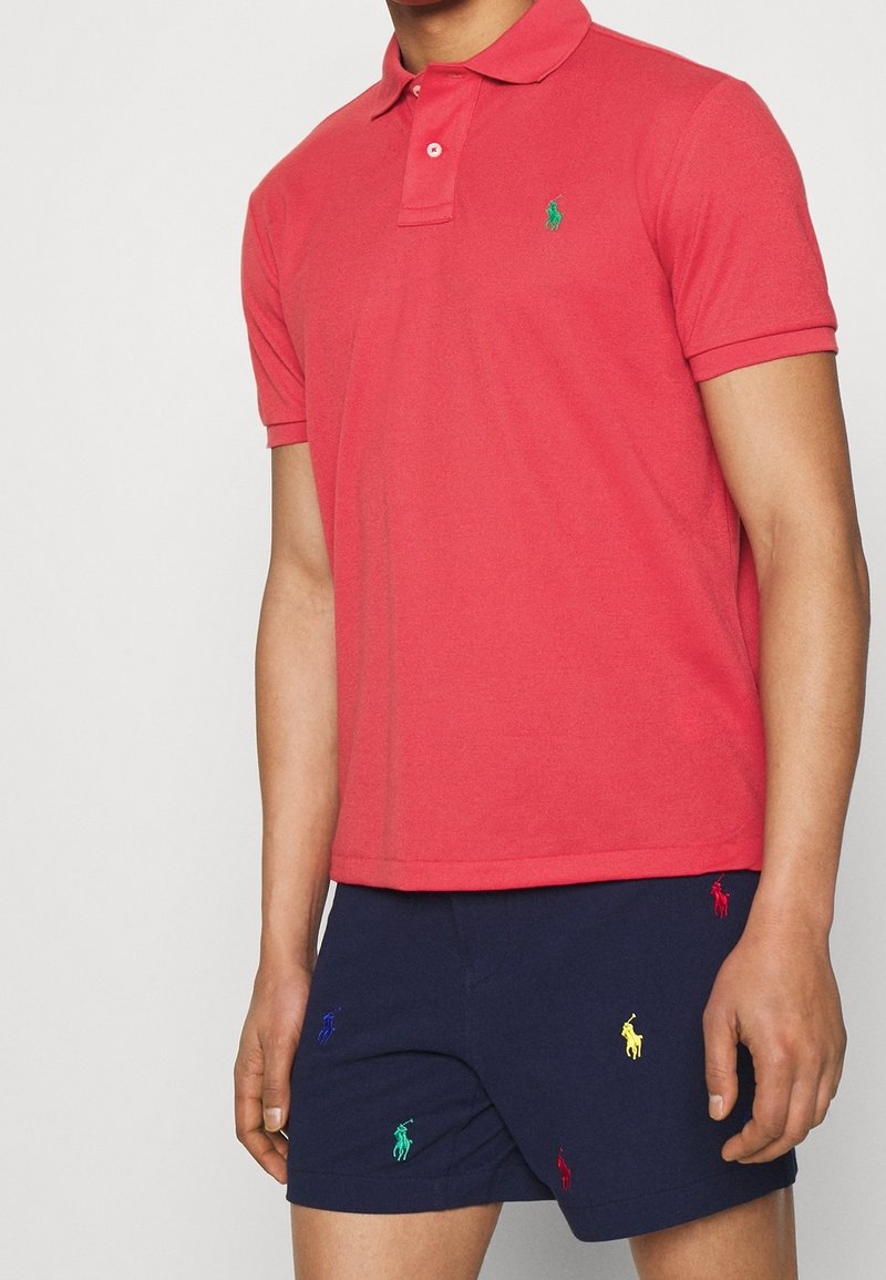 Homme portant un polo rouge à manches courtes et un short bleu marine avec de petits logos brodés multicolores.