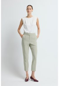 Vrouw die staat met haar handen in de zakken, gekleed in een mouwloze witte blouse, lichtgrijze op maat gemaakte broek en bordeauxkleurige hakken tegen een witte achtergrond.