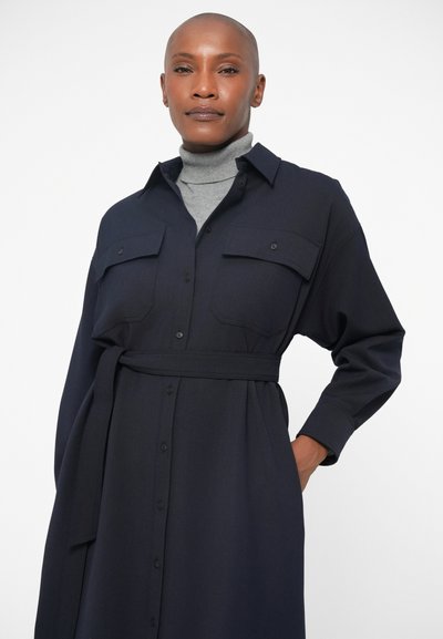 Ναυτικό μπλε φόρεμα με μακριά μανίκια, μπροστινό κουμπί και δύο τσέπες στο στήθος. Το ύφασμα φαίνεται απαλό, σχεδιασμένο με μια ταιριαστή ζώνη στη μέση.