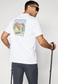Camiseta blanca de algodón con un gráfico colorido de montañas y bosques en la parte posterior. Pantalones deportivos grises y bastones de senderismo negros completan el atuendo.
