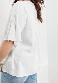 Femme portant une blouse blanche ample avec des détails plissés sur les manches et le dos, associée à un jean en denim bleu clair.