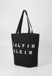 Juodas kanvas pirkinių krepšys su dviem rankenomis ir ryškiomis baltomis raidėmis, kuriomis parašyta "CALVIN KLEIN" ant priekio. Struktūruota forma ir lygi tekstūra.