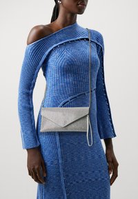 Silver mesh clutch bag with an envelope shape, worn crossbody. The textured blue ridged dress features an off-shoulder design and button accents.