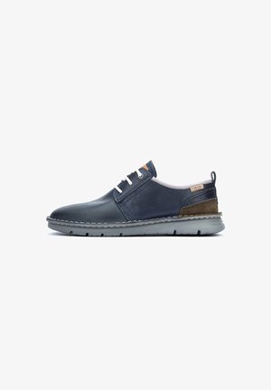 Zapato casual de cuero azul marino con cordones blancos, acento en tacón de ante marrón y suela de goma gris, vista lateral sobre fondo blanco.