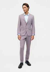 Moški, oblečen v svetlo vijolično obleko s svetlo modro srajco in črnimi poslovnimi čevlji, stoji pred enobarvno belo steno.