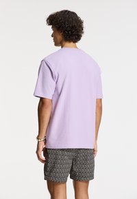 Levandų spalvos trumpomis rankovėmis marškinėliai iš medvilnės, apvaliu iškirpimu, laisvo kirpimo. Suderinti su juodais šortais su geometriniu raštu.