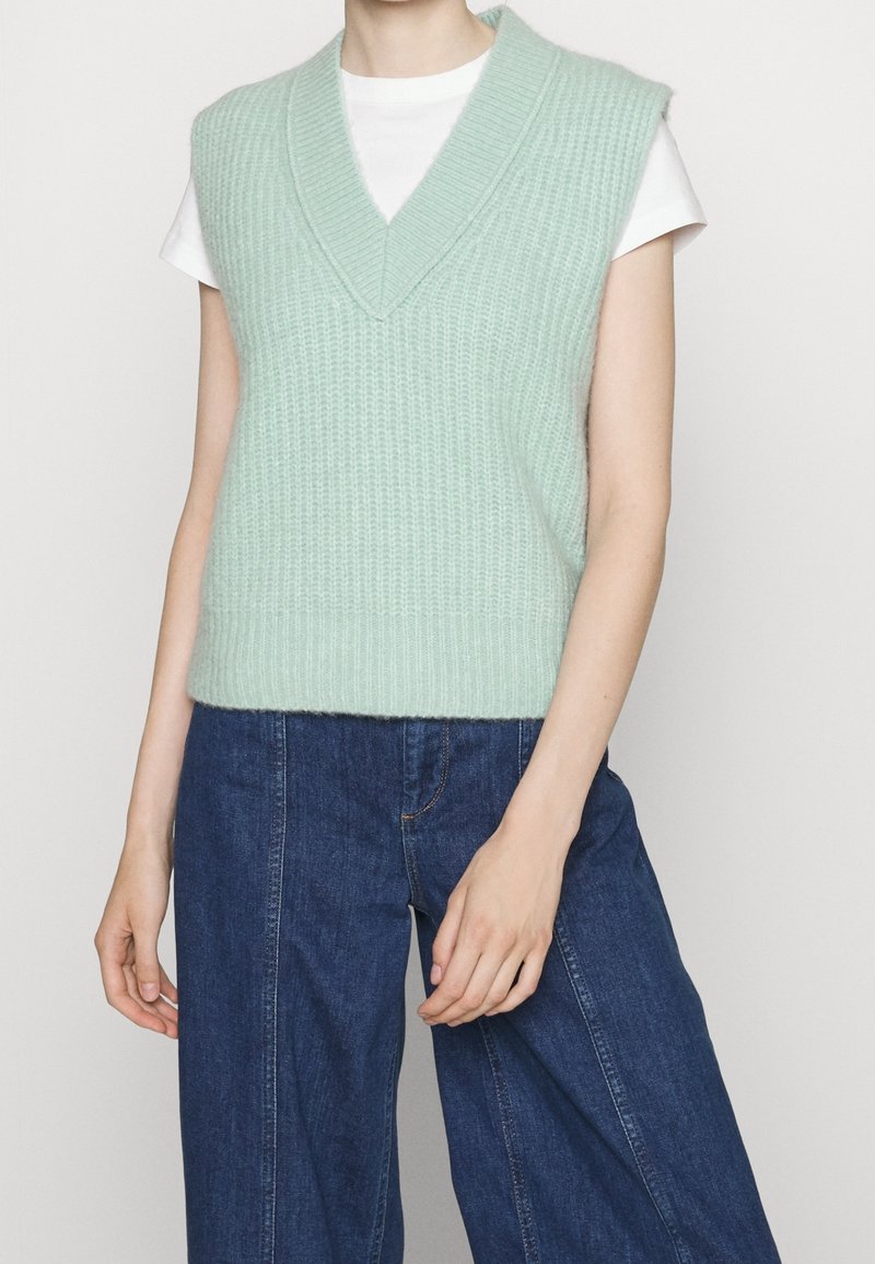 Mintgroene gebreide mouwloze sweater met een V-hals en geribbeld textuur, gedragen over een wit T-shirt. Gepaard met donkerblauwe wijde jeans.