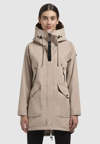 Giacca beige con colletto alto e cappuccio regolabile, dotata di due tasche frontali, chiusura con zip e orlo con cordino. Etichetta del marchio nera visibile.