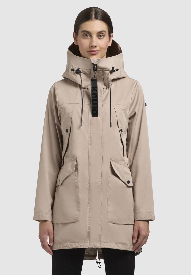Giacca beige con colletto alto e cappuccio regolabile, dotata di due tasche frontali, chiusura con zip e orlo con cordino. Etichetta del marchio nera visibile.