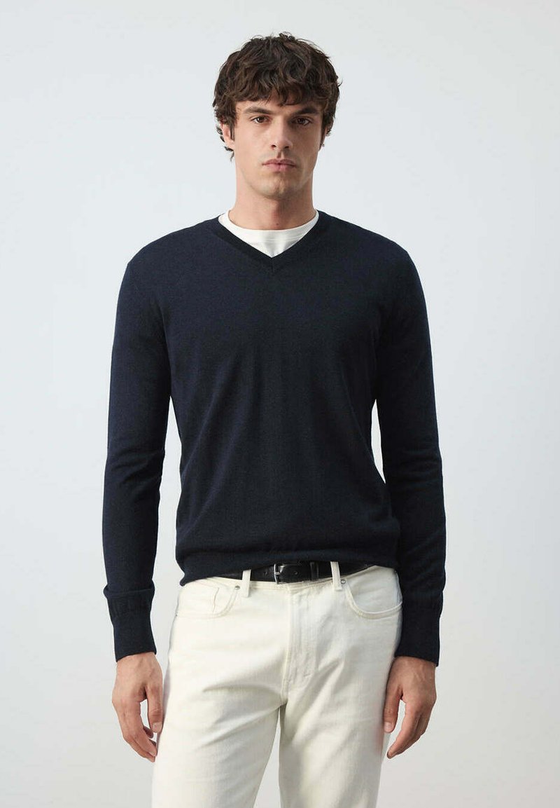 Homme aux cheveux courts et bouclés portant un pull noir en V par-dessus une chemise blanche et un pantalon blanc, debout devant un fond clair uni.