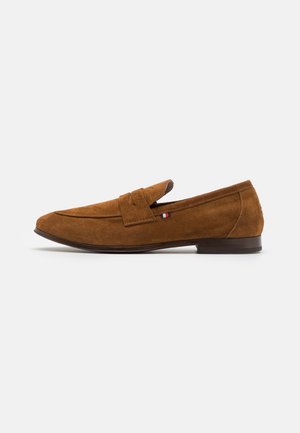 Mocassins pour hommes en daim marron avec semelle plate, couture moc toe et petit écusson en tissu rouge, blanc et bleu sur le côté.