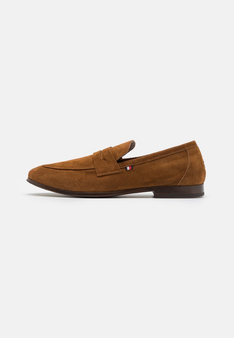 Mocassins pour hommes en daim marron avec semelle plate, couture moc toe et petit écusson en tissu rouge, blanc et bleu sur le côté.