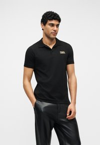 Άνδρας που φορεί μαύρο κοντομάνικο πόλο με το λογότυπο "KARL LAGERFELD", συνδυασμένο με μαύρο δερμάτινο παντελόνι, στέκεται σε λευκό φόντο.