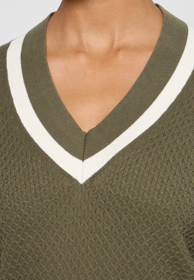 Pullover en tricot vert olive avec un motif texturé, présentant un col en V et une bordure rayée blanche le long des bords.