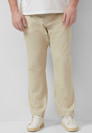 Mężczyzna ubrany w beżowe spodnie o prostych nogawkach, białe trampki i białą koszulkę na jednolitym jasnym tle.