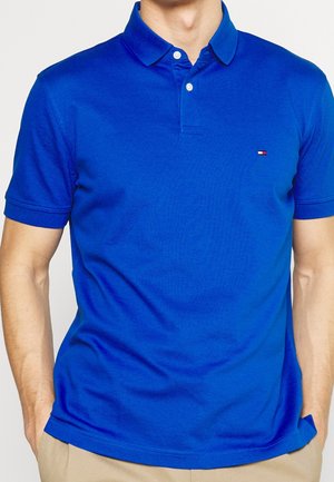 Homme portant un polo bleu vif à manches courtes avec deux boutons blancs et un petit logo rectangulaire sur la poitrine.