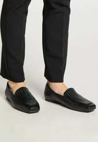 Zwarte loafers van gestructureerd leer, met een gevlochten patroon, vierkante neus en een platte zool, gecombineerd met zwarte op maat gemaakte broek.