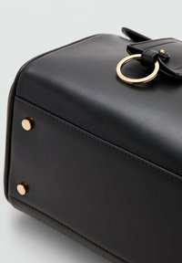 Bolso de cuero negro con forma rectangular, que presenta un herraje de anillo metálico dorado y tachuelas doradas en la base. Textura suave con detalles de costuras.