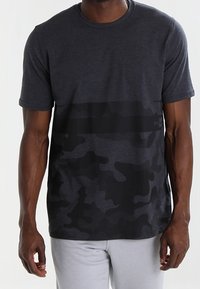 Camiseta gris de manga corta con un patrón de camuflaje negro en el torso, confeccionada en tela suave, con cuello redondo y corte relajado.