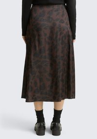 Femme portant une jupe midi à motifs marron foncé et noirs, des chaussettes noires et des chaussures noires, se tenant devant un fond clair et uni.