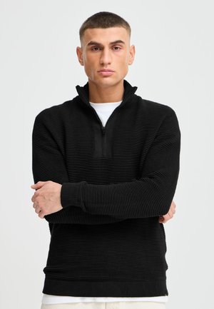 Junger Mann mit kurzen Haaren, der einen schwarzen, texturierten Pullover mit Viertel-Reißverschluss trägt und die Arme vor einem schlichten hellen Hintergrund verschränkt hat.