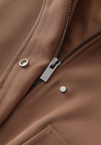 Bruine stof met diagonale ribbels, voorzien van een zilveren rits en drukknoop. Rits gedeeltelijk geopend, waardoor de soepele binnenvoering zichtbaar is.