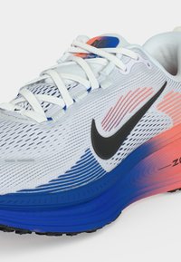 Λευκό αθλητικό παπούτσι με μαύρο λογότυπο Nike, σόλα με μπλε και πορτοκαλί διαβαθμισμένο χρώμα, λευκά κορδόνια και επάνω μέρος από δίχτυ για διαπνοή.
