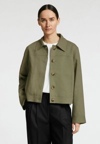 Chaqueta corta de color verde oliva, hecha de un tejido resistente, con cuello puntiagudo, cinco grandes botones marrones y bolsillos laterales.
