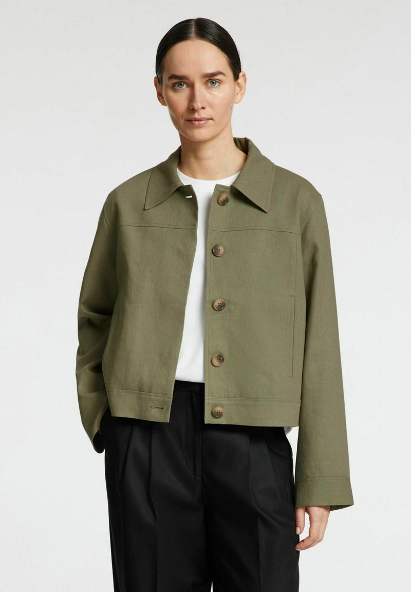 Chaqueta corta de color verde oliva, hecha de un tejido resistente, con cuello puntiagudo, cinco grandes botones marrones y bolsillos laterales.
