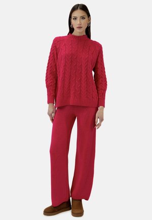 Kobieta ubrana w czerwony sweter w warkocze i pasujące szerokie spodnie, brązowe buty wsuwane, stoi na białym tle.