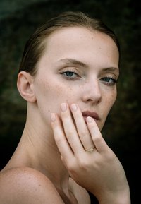 Zlatý prsteň s kvetinovým dizajnom, ktorý obsahuje centrálny kameň a menšie akcenty. Modelka má svetlú pokožku, svetlohnedé vlasy a prirodzené nechty.