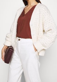 Cardigan en tricot crème avec un motif texturé, associé à un t-shirt en V couleur rouille, un pantalon blanc et un sac à main bordeaux. Boutons du cardigan visibles.