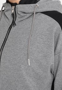 Sweatshirt gris avec capuche, présentant des accents noirs aux épaules et une fermeture éclair centrale. Texture lisse avec cordons de serrage réglables.