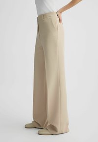 Pantaloni beige a gamba larga con una texture liscia, vita alta, tasche laterali e un bottone dorato come dettaglio. Abbinati a calzature di colore chiaro.