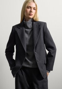 Blazer de lana gris oscuro con hombros estructurados, solapas de pico y dos botones. Combinado con un suéter de cuello alto acanalado gris claro debajo.