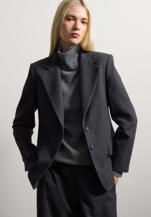 Blazer en laine gris foncé avec des épaules structurées, des revers en pointe et deux boutons. Associé à un pull col roulé côtelé gris clair en dessous.