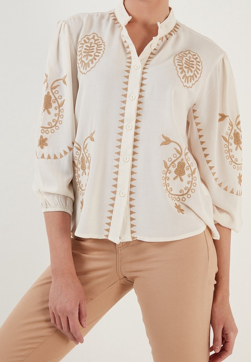Blouse blanche boutonnée avec broderie florale beige sur la poitrine et les manches, portée avec un pantalon beige.